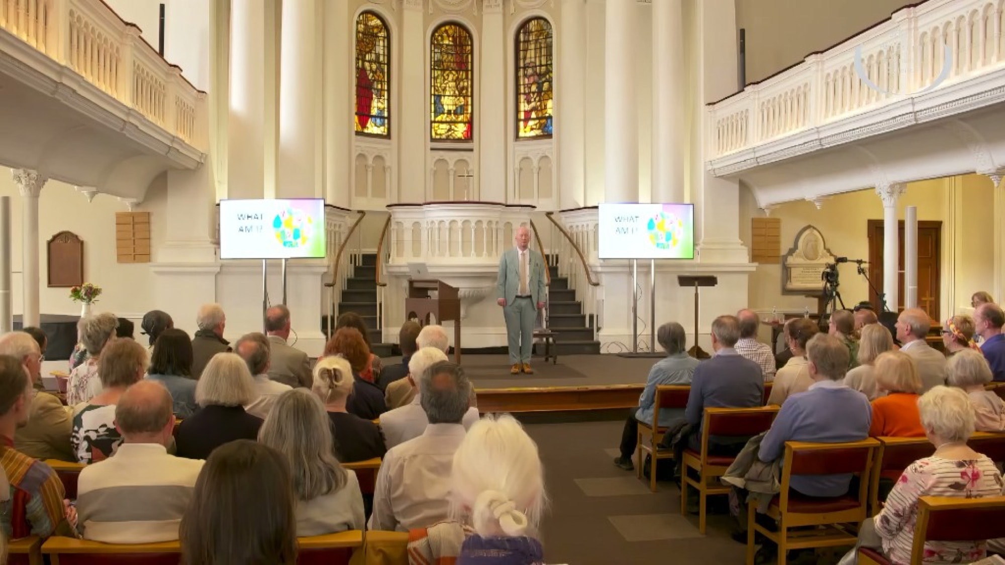 Public lecture in a bright hall: speaker at the front, audience seated, screens reading What am I?