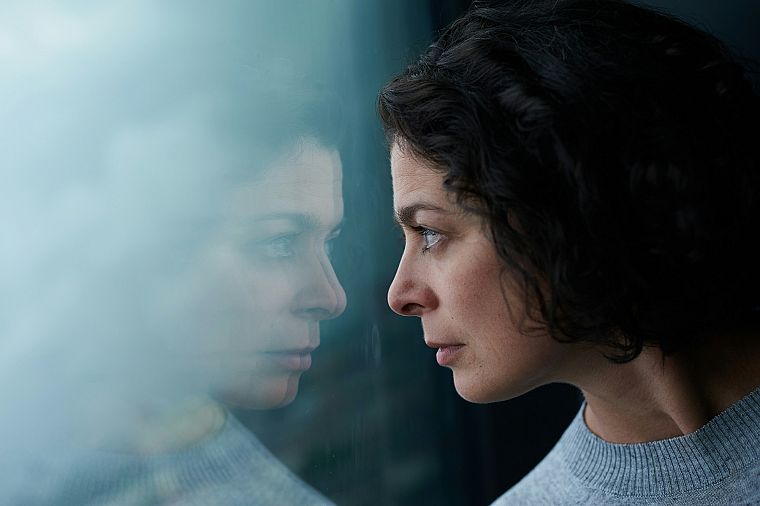 A woman looking through a window, her face reflected in the glass