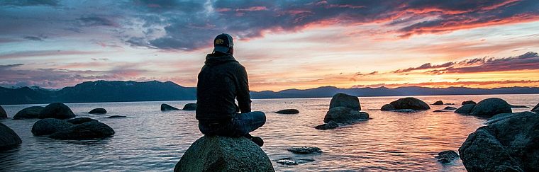 A person sitting on a rock at the water's edge, watching the sunset