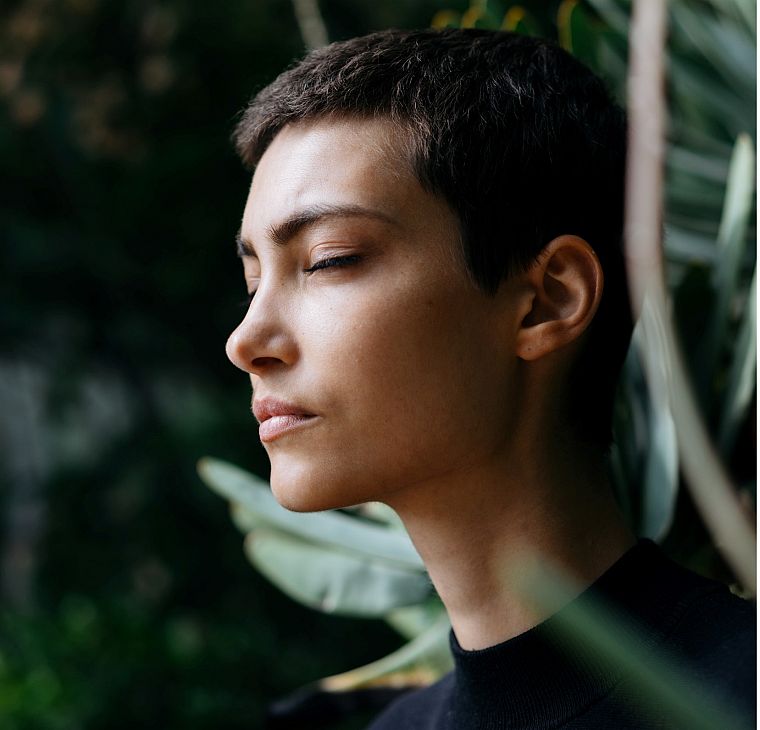 A person with their eyes closed, standing among leaves in soft daylight